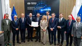Representatives of partner institutions pose for a group photograph during the signing of a cooperation agreement, holding the signed document in front of a display screen and national and institutional flags.
