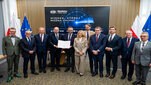 Representatives of partner institutions pose for a group photograph during the signing of a cooperation agreement, holding the signed document in front of a display screen and national and institutional flags.