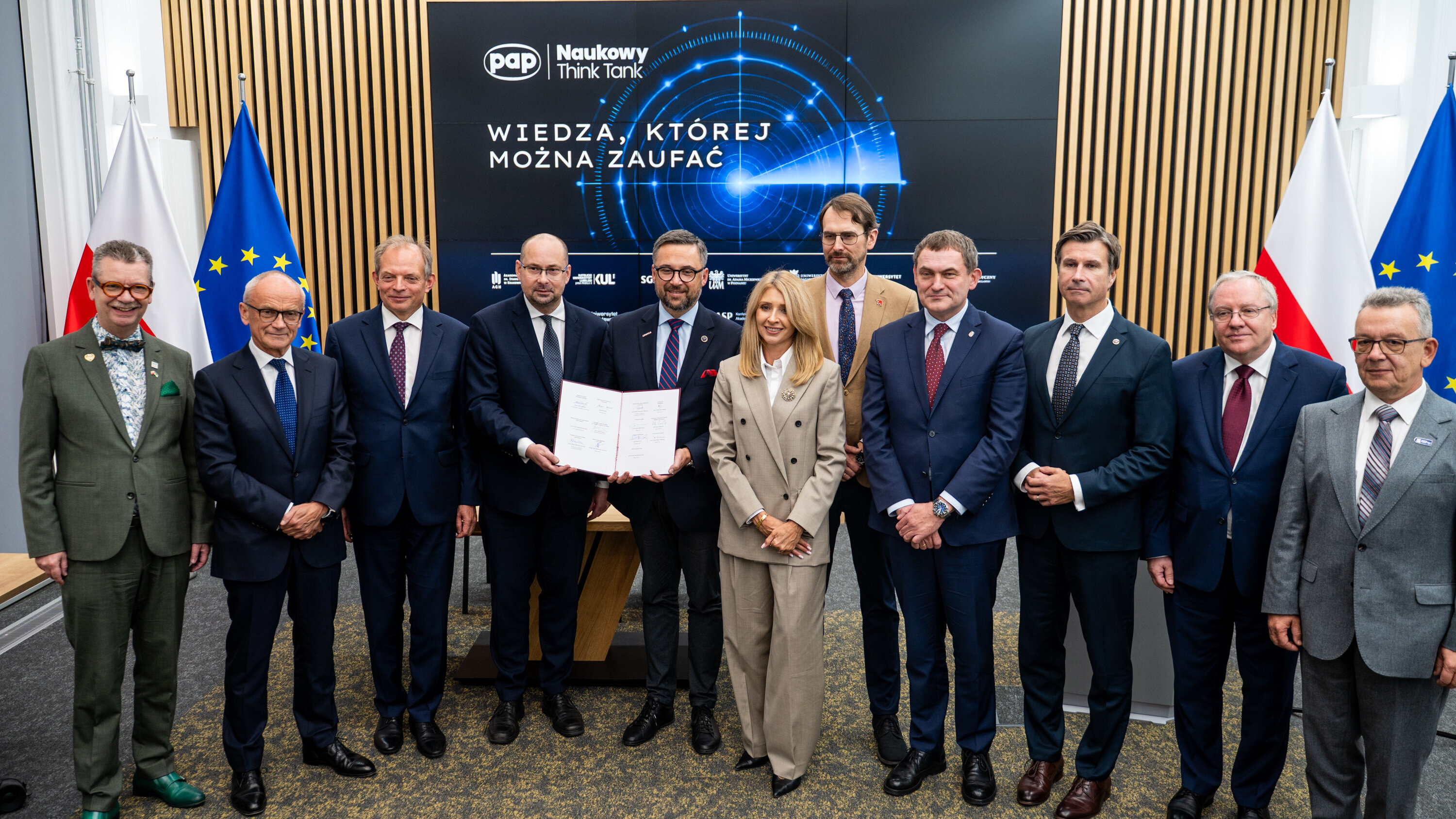 Representatives of partner institutions pose for a group photograph during the signing of a cooperation agreement, holding the signed document in front of a display screen and national and institutional flags.