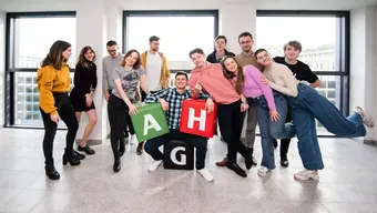 Grupa studentów i studentek AGH pozuje do zdjęcia w jednym z budynków uczelni. Elementem kompozycyjnym ujęcia są gadżety promocyjne w postaci kolorowych pufy z literami „A”, „G” i „H”.