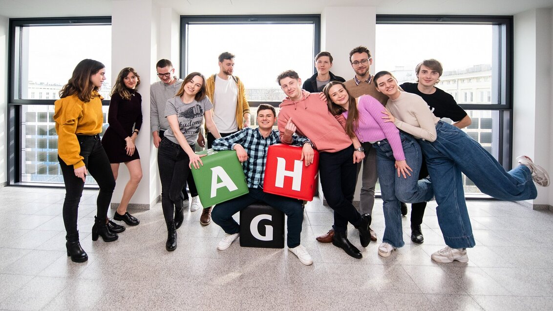 Grupa studentów i studentek AGH pozuje do zdjęcia w jednym z budynków uczelni. Elementem kompozycyjnym ujęcia są gadżety promocyjne w postaci kolorowych pufy z literami „A”, „G” i „H”.