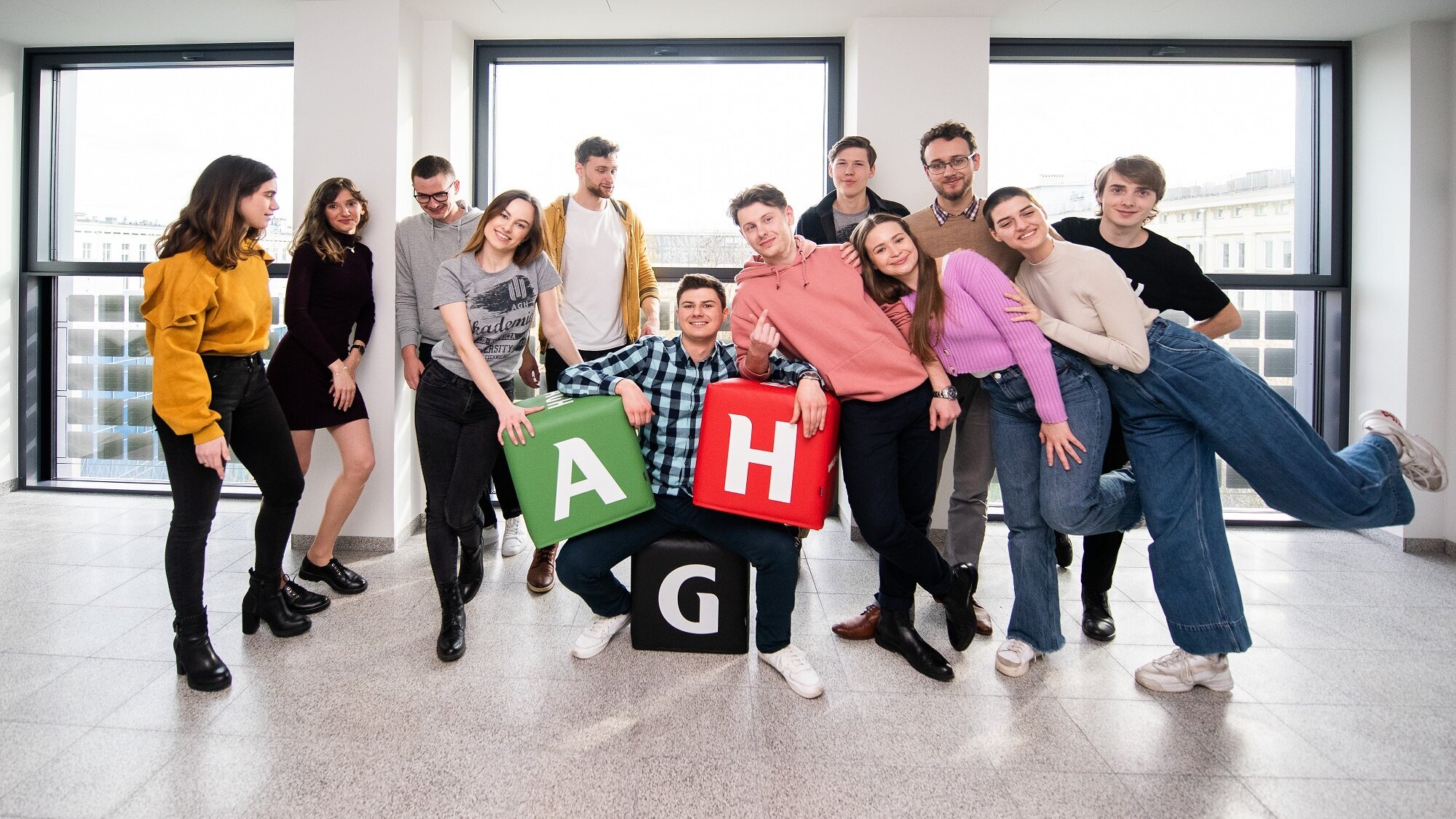 Grupa studentów i studentek AGH pozuje do zdjęcia w jednym z budynków uczelni. Elementem kompozycyjnym ujęcia są gadżety promocyjne w postaci kolorowych pufy z literami „A”, „G” i „H”.