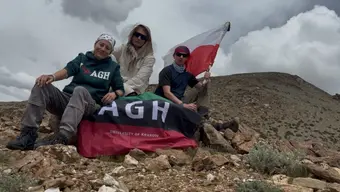 Trzy osoby siedzą na skalistym terenie z górskim krajobrazem w tle. Trzymają flagę Polski oraz flagę z napisem „AGH University of Krakow”.