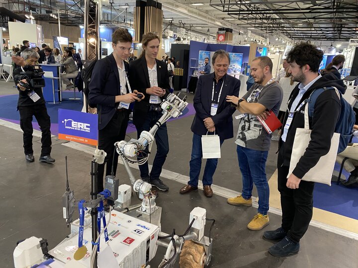 A huge exhibition hall. The exhibitors present their projects and innovations at their booths. In the foreground, the Kalman rover with a group of five. One of them talks to the others, probably describing the robot. In the background, there are numerous booths and groups of people around them.