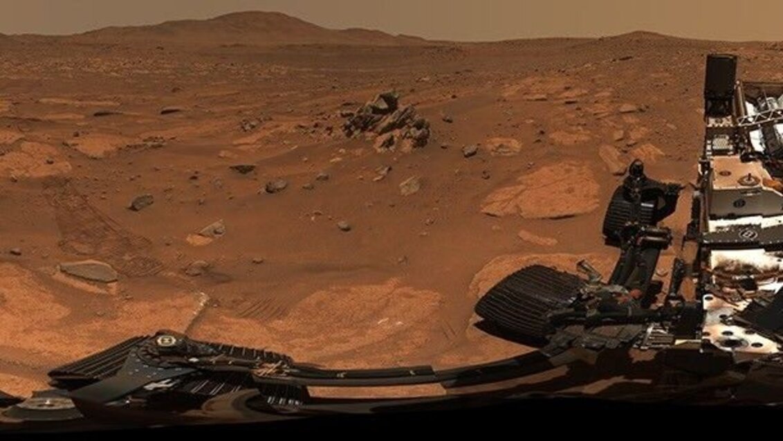 A panoramic image of the Martian surface taken by a rover, featuring reddish-brown rocky terrain with distant hills under a dusty sky.