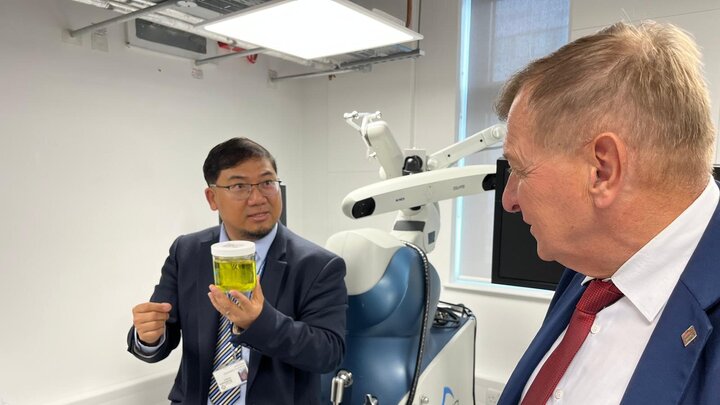 Two elegant men in laboratory setting. One of them is holding a clear jar containing yellow fluid, explaining something, indicating with his hand.