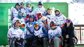 A group photo of the AGH UST snowboarding team (15 people). All wearing sports clothing, helmets, and goggles. Their faces express joy, they are smiling.