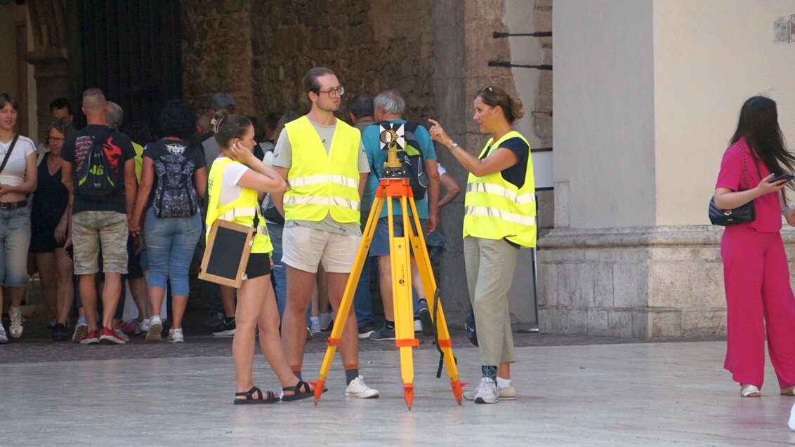 Three people in high-visibility vests standing near a geodetic tripod with tourists in the background.