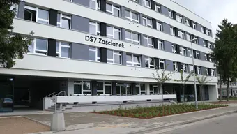 A modern four-story student hall of residence named "DS7 Zaścianek" with a white and grey facade and grid-like window arrangement. The entrance features stairs and a ramp for accessibility. In front, there's a landscaped area with trees and a paved sidewalk.
