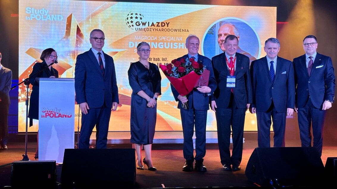 Image of the winner of a special award at a gala; a few people standing on a stage, one man holding flowers and an award.