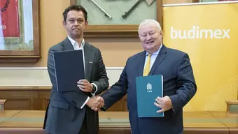 Two formally dressed men shaking hands while holding documents. Behind them, there is a yellow banner with the name "budimex"