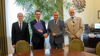 Four men standing next to each other in a row. The two in the middle shake hands.