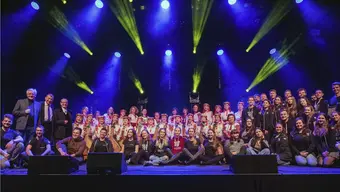 Grupowe zdjęcie członków zespołu (siedzą) oraz osób z widowni (siedzą i stoją). Po lewej stronie stoją Wiesław Ochman i prof. Jerzy Lis.