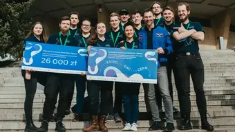 Grupowe zdjęcie kilkunastuosobowego zespołu AGH Space Systems razem ze Sławoszem Uznańskim. Osoby na zdjęciu są uśmiechnięte, stoją obok siebie na schodach, przed budynkiem. W dłoniach trzymają plansze z informacją o nagrodzie oraz konkursie.