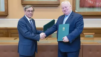 Na zdjęciu sygnatariusze umowy tuż po jej podpisaniu podają sobie dłonie w uścisku. Zdjęcie wykonane w sali konferencyjnej.