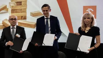 Image of three people, two men and a woman, holding opened folders with a signed document in an official setting, standing against a backdrop with jenga blocks and the logo of Orlen