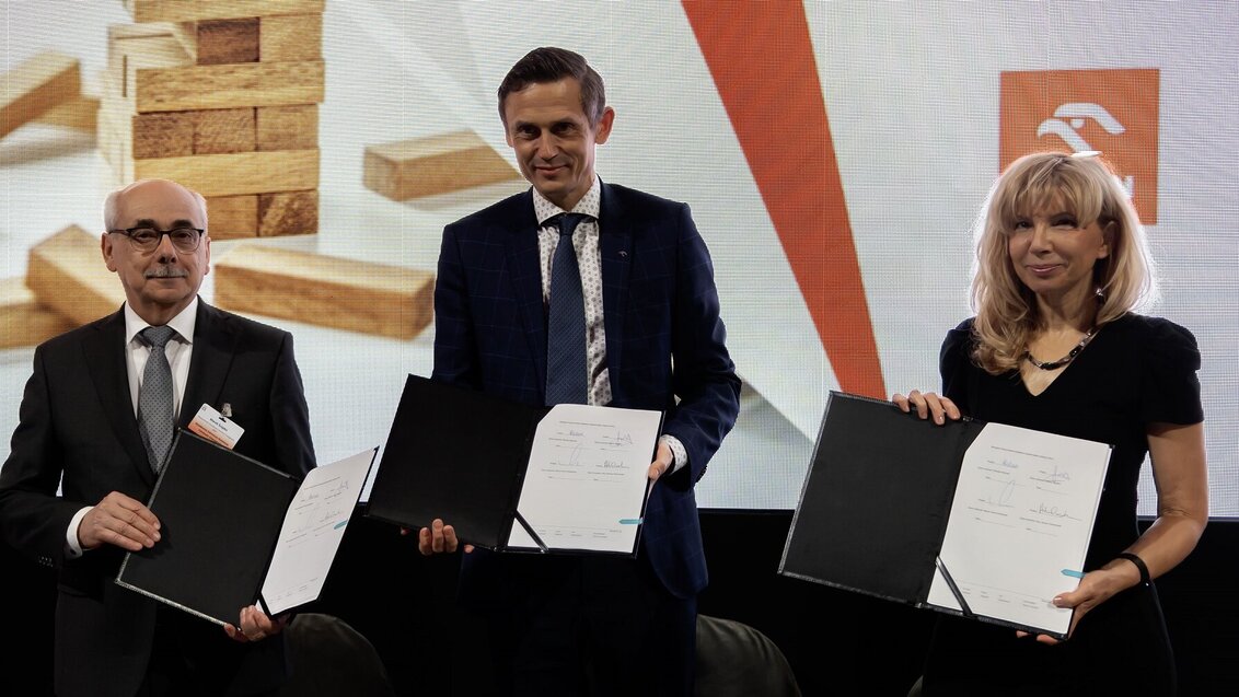 Image of three people, two men and a woman, holding opened folders with a signed document in an official setting, standing against a backdrop with jenga blocks and the logo of Orlen