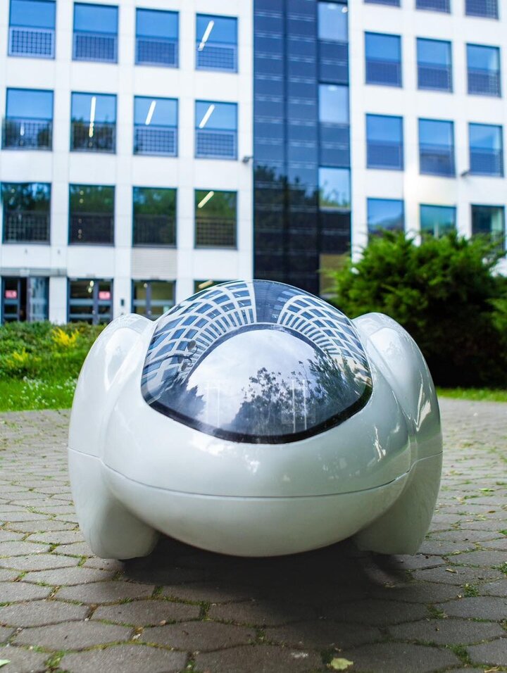 The image features a sleek, reflective dome-like car outdoors, surrounded by a modern building and greenery.