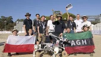 Image of the AGH Space Systems team with their rover Kalman in front of them. Some of the students are holding a Polish flag and an AGH University banner