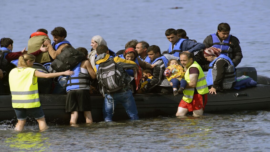 The photo shows a rubber raft with refugees from the Middle East. Next to it in shallow water, there are two people wearing high-visibility vests. One of them is holding a baby in his hands.