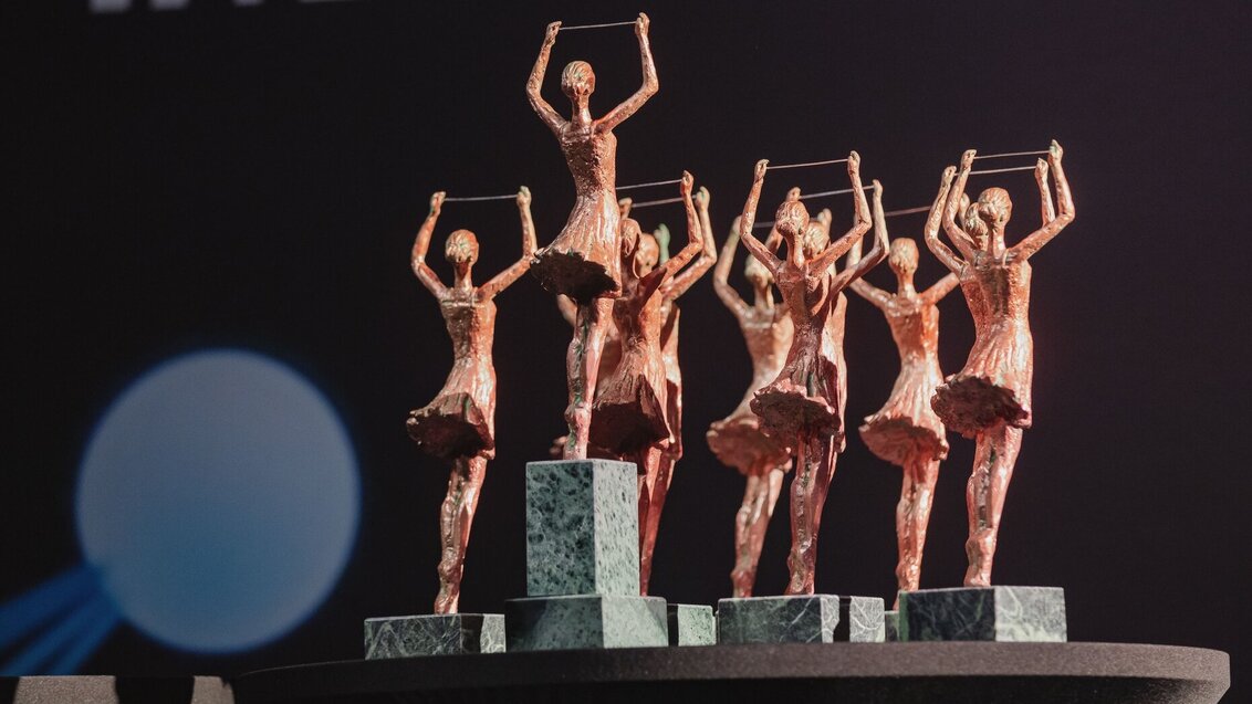 A coloured photo of commemorative statuettes presented to the winners of the competition. The statuettes represent female figures in floaty dresses with hands reaching up and holding a string.