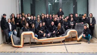 Grupa kilkudziesięciu osób stoi lub siedzi wewnątrz dużego pomieszczenia warsztatowego. Zespół pozuje wokół dużego, podłużnego elementu konstrukcyjnego o nieregularnym kształcie, ustawionego na podłodze przed grupą. W tle znajdują się wysokie okna z metalowymi roletami oraz surowe, industrialne wnętrze.