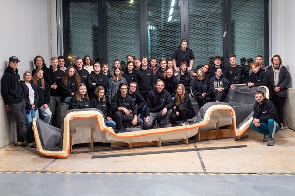 Grupa kilkudziesięciu osób stoi lub siedzi wewnątrz dużego pomieszczenia warsztatowego. Zespół pozuje wokół dużego, podłużnego elementu konstrukcyjnego o nieregularnym kształcie, ustawionego na podłodze przed grupą. W tle znajdują się wysokie okna z metalowymi roletami oraz surowe, industrialne wnętrze.