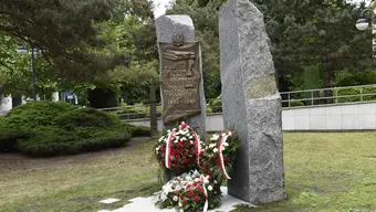 This is a memorial monument with a bronze plaque, wreaths, and red-and-white ribbons in a park setting.