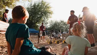 Two kids sitting on the ground resembling the surface of Mars, watching a rover challenge surrounded by people