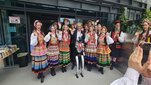 Zdjęcie grupowe członków i członkiń zespołu "Krakus" w strojach regionalnych oraz osoby w ciemnym garniturze. Przed grupą stoi humanoidalny robot z wiszącymi na korpusie czerwonymi koralami. Wszyscy stoją przed wejściem do budynku Biblioteki Głównej.
