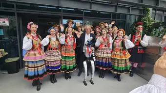 Zdjęcie grupowe członków i członkiń zespołu "Krakus" w strojach regionalnych oraz osoby w ciemnym garniturze. Przed grupą stoi humanoidalny robot z wiszącymi na korpusie czerwonymi koralami. Wszyscy stoją przed wejściem do budynku Biblioteki Głównej.