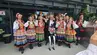 Zdjęcie grupowe członków i członkiń zespołu "Krakus" w strojach regionalnych oraz osoby w ciemnym garniturze. Przed grupą stoi humanoidalny robot z wiszącymi na korpusie czerwonymi koralami. Wszyscy stoją przed wejściem do budynku Biblioteki Głównej.