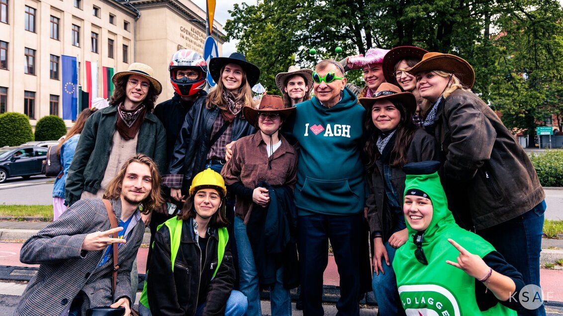 AGH University Rector in an AGH University sweatshirt posing for a photograph with a group of students wearing various costumes. The main building of AGH University is visible in the background.