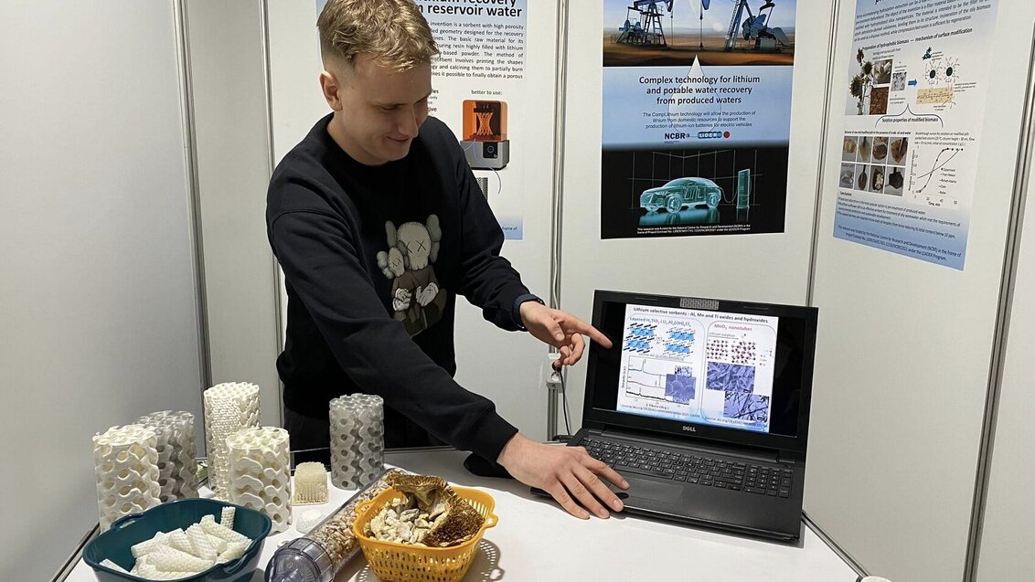 Image of Igor Danowski with 3D-printed solutions for lithium recovery standing behind a desk and pointing his finger at a laptop screen with scientific information behind these solutions