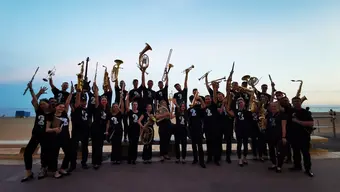 A large group of musicians dressed in black, holding up all kinds of brass instruments. A beach and sea in the background.