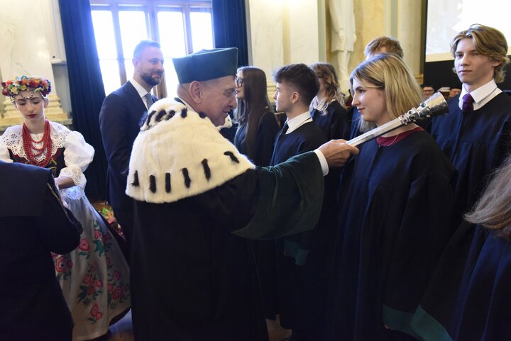 The Rector dressed in his academic robes placing his sceptre on a student's shoulder.