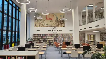 Zdjęcie czytelni po remoncie: jasna, nowoczesna przestrzeń z rzędami biurek, otoczona regałami pełnymi książek; na ścianie centralnej duży zegar oraz dekoracyjny mural z motywami naukowymi.