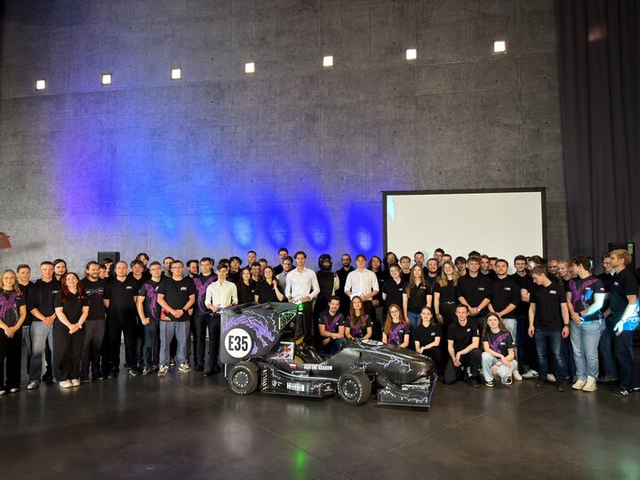 A group of several dozen people gathered around the racing car in a large industrial hall.