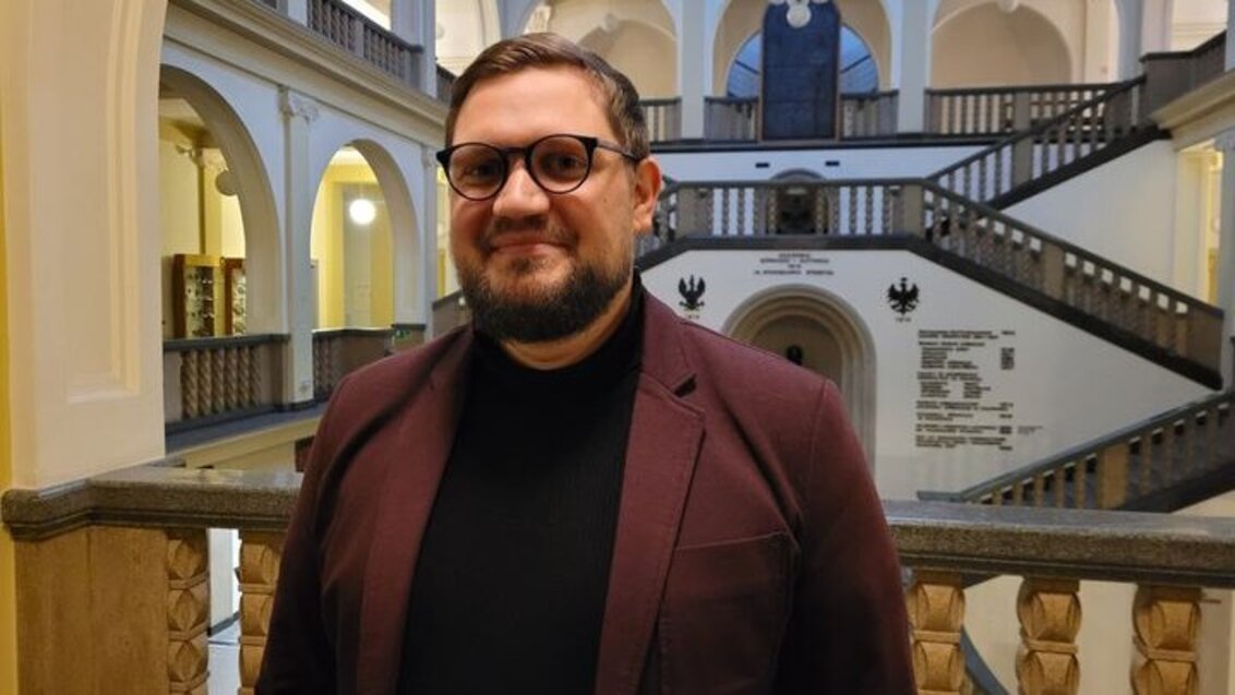 A smiling man with glasses wearing a burgundy jacket and a black polo neck. The photo was taken in the main hall of the Main Building, with a pillared cloister and stairs in the background.