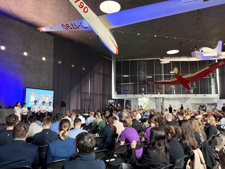A large hall where the audience is gathered. Aircraft suspended from the ceiling, forming part of the museum’s exhibition, are visible.