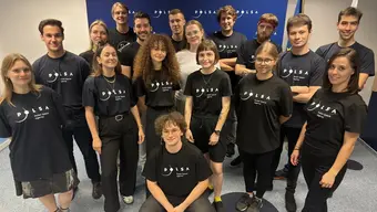 Grupowe zdjęcie - kilkanaście młodych osób (kobiet i mężczyzn) pozuje do zdjęcia. Większość z nich ma na sobie czarne koszulki z białym napisem POLSA Polish Space Agency.