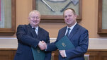 Two elderly men pose for a photo, shaking each other’s hands. Both are wearing suits. Both hold green document folders with gold AGH logos thereon. In the background, there are showcases with the rector’s insignia on display.