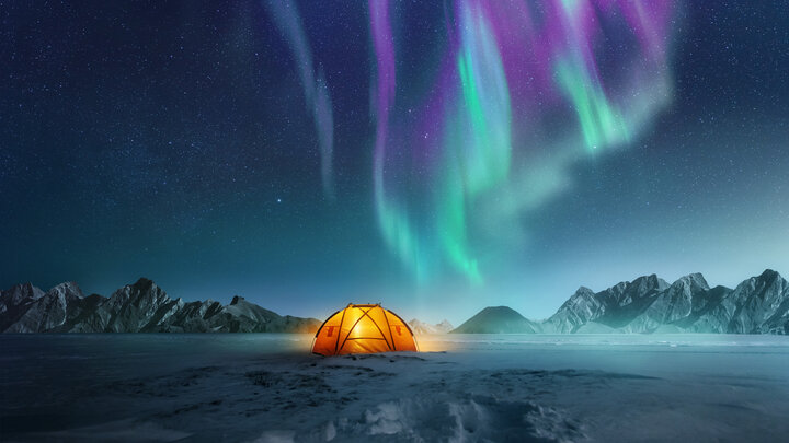 Zdjęcie pejzażu arktycznego nocą. Na pierwszym planie podświetlony od środka namiot. W tle niebo z zorzą polarną. Na horyzoncie pasmo górskie.