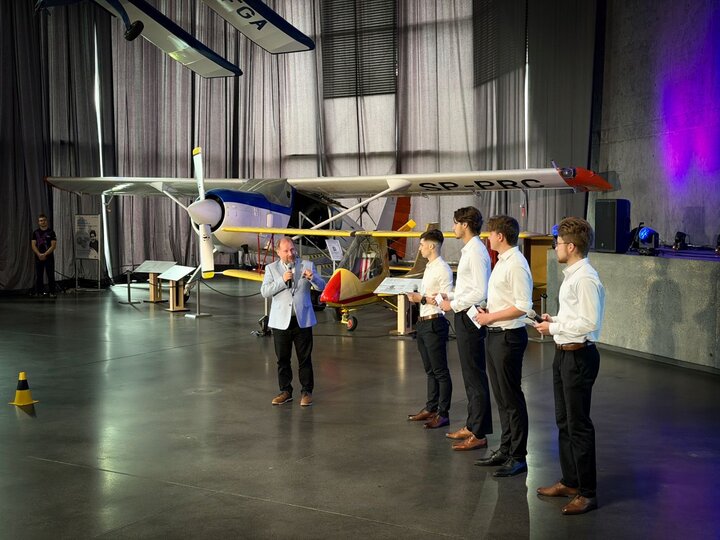 An elegant man is speaking at the ceremony, flanked by four members of the AGH Racing team, with an aeroplane – part of the museum’s exhibition – visible in the background.