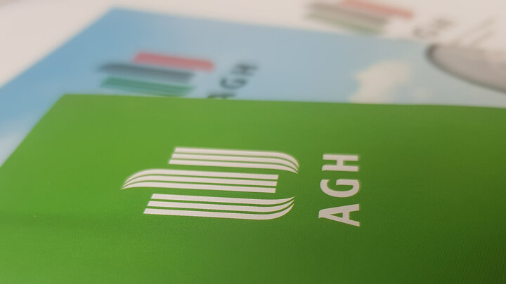Zdjęcie makro materiałów poligraficznych. Na pierwszym planie zielona kartka z nadrukowanym białym logotypem AGH.
