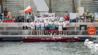 A large group of people posing for a photo in a marina, a red boat is floating in front of them. One person in the group is waving a Polish flag, and three other people are holding an AGH University banner.