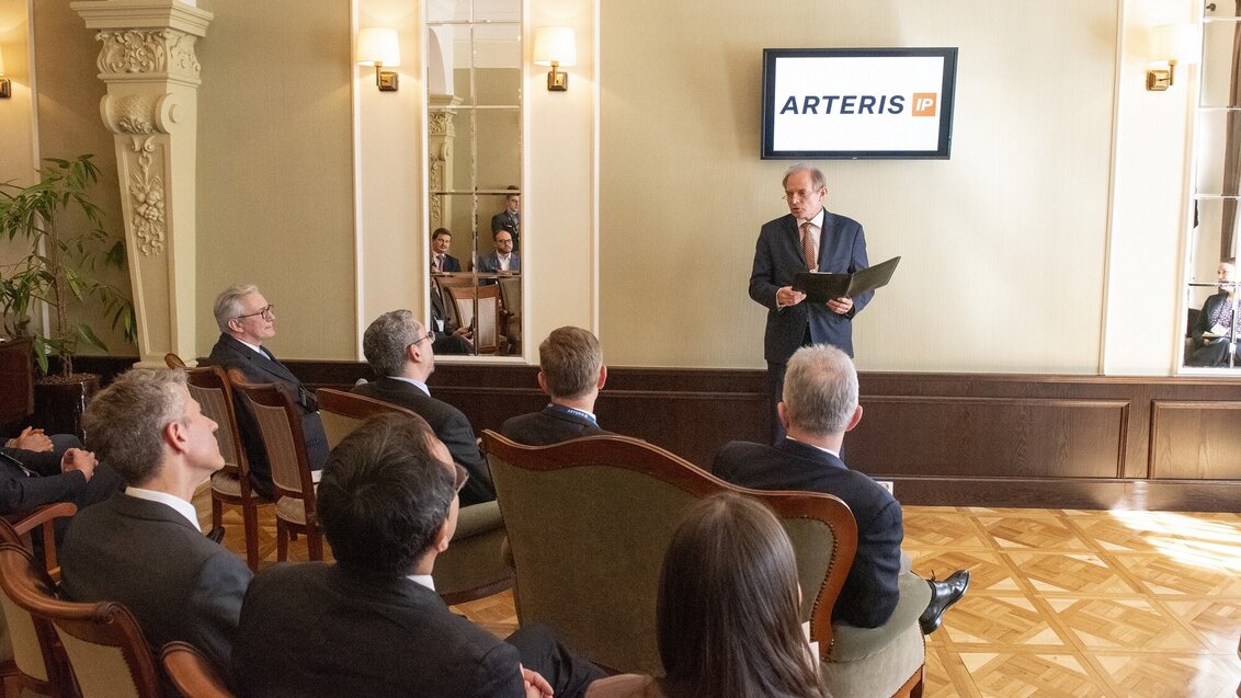 An image of Professor Gorgoń holding a folder, speaking to a group seated in an elegant room, with a screen behind him displaying the logo og Arteris
