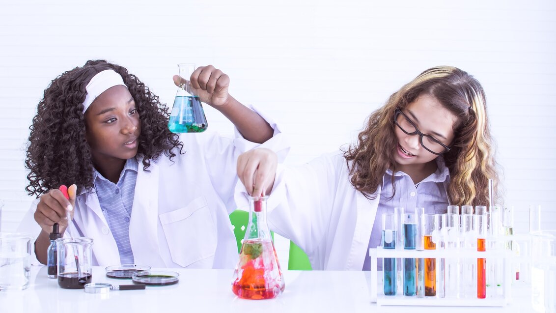 Two young girls in lab coats working with beakers and vials filled with colourful liquids.