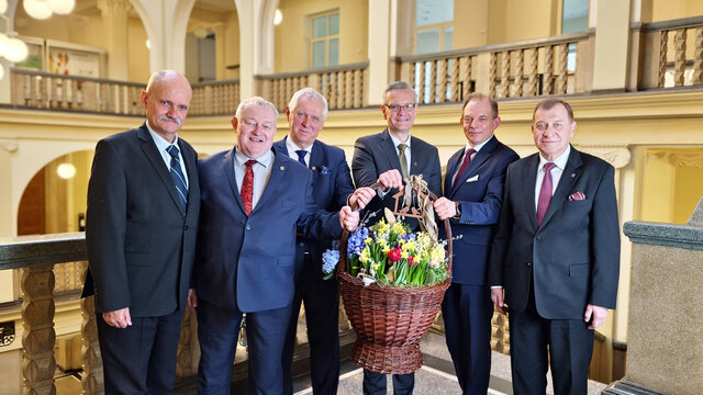 Zdjęcie przedstawia sześciu uśmiechniętych mężczyzn w średnim wieku w biznesowych strojach skierowanych w stronę widza. Czterech z nich trzyma wielki kosz z kwiatami oraz wielkanocnymi dekoracjami. 