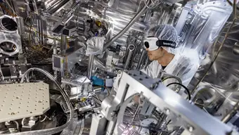 Na zdjęciu nowoczesne laboratorium ze specjalistycznym sprzętem, w którym naukowiec (ubrany w fartuch oraz ochronne rękawice, czepek i okulary) przeprowadza doświadczenie.